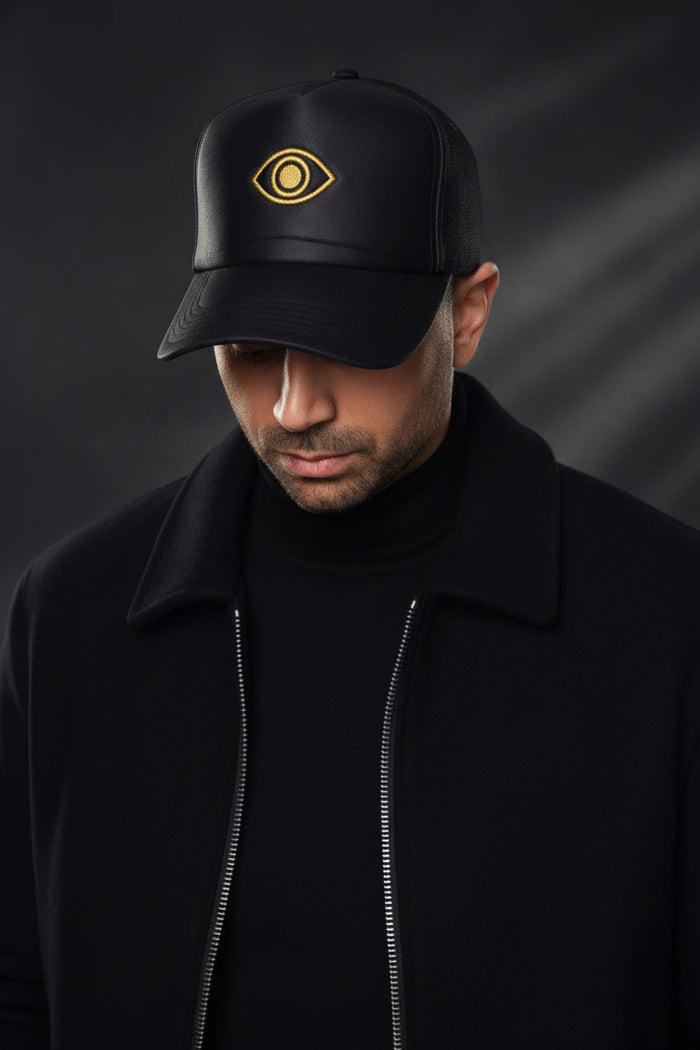 Person wearing a black cap with a gold eye emblem on a gray background
