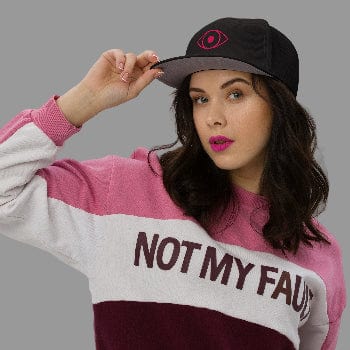 Woman wearing a pink and white sweatshirt with 'NOT MY FAULT' text, holding a black cap.
