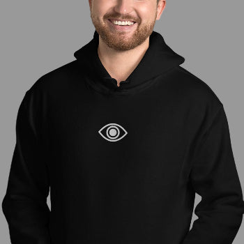 Man wearing a black hoodie with an eye emblem against a white brick wall.